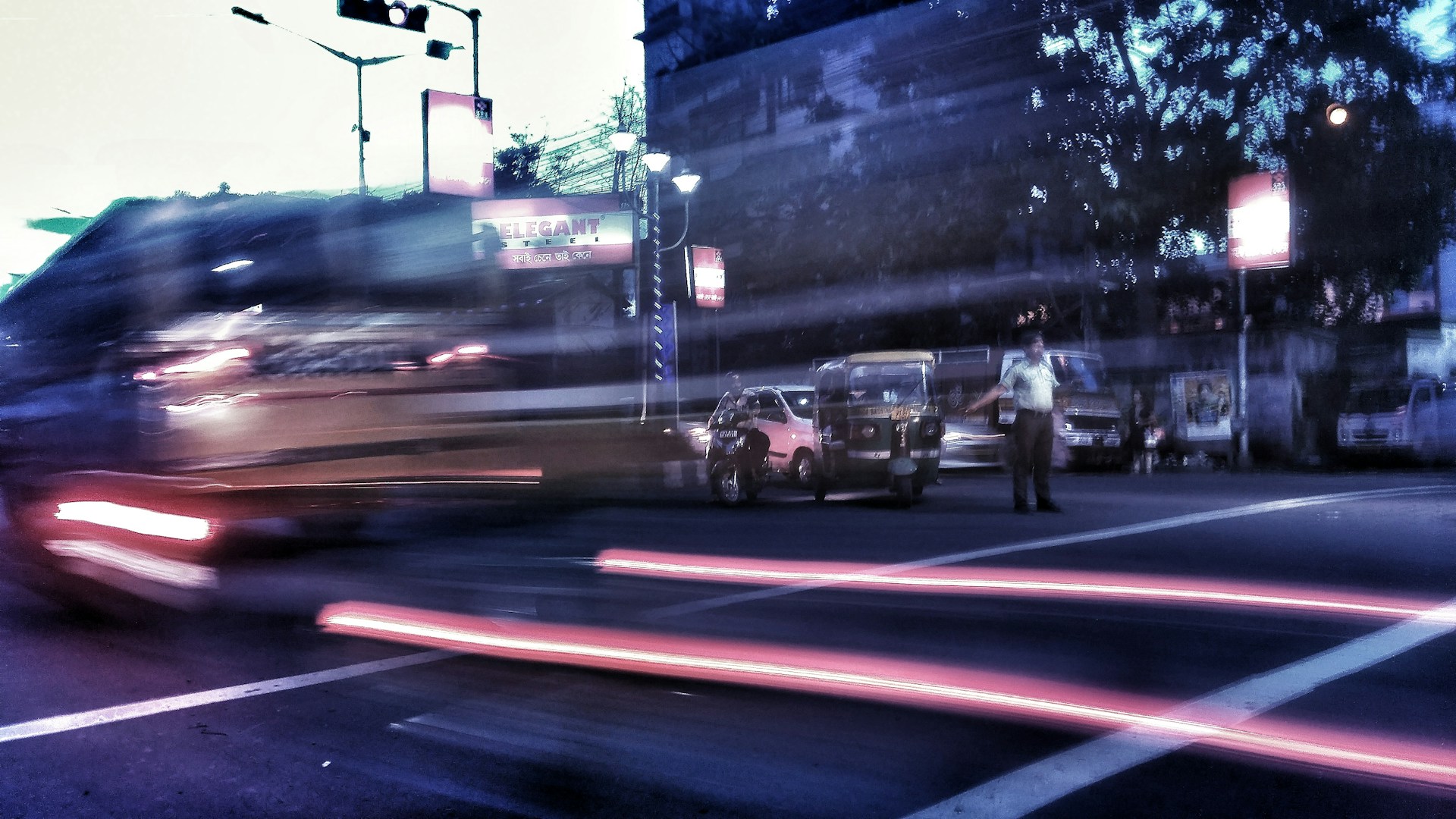 Motion blur effect captures fast-moving vehicles at a city intersection during the evening. Streetlights cast a blue and reddish hue over the scene, with an auto rickshaw and a figure at the side of the road.