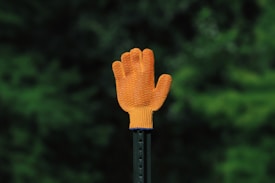 A bright orange glove is positioned upright on a dark pole against a blurry background of lush green foliage. The texture of the glove is visible, highlighting its knitted material.