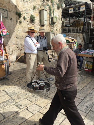 A street scene in an old city with two men wearing hats and cameras across their necks standing and observing. In the foreground, a man is carrying a tray filled with cups. The background features stone walls with some greenery, electrical boxes, and signage. There are also market stalls with various items.