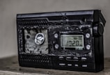 A damaged electronic device with a digital display showing the frequency 102.0 MHz. The surface of the device appears melted or burnt, with a rough texture around the front panel. The right side has several control buttons labeled with functions such as On/Off and Light.