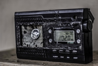 A damaged electronic device with a digital display showing the frequency 102.0 MHz. The surface of the device appears melted or burnt, with a rough texture around the front panel. The right side has several control buttons labeled with functions such as On/Off and Light.