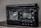 A damaged electronic device with a digital display showing the frequency 102.0 MHz. The surface of the device appears melted or burnt, with a rough texture around the front panel. The right side has several control buttons labeled with functions such as On/Off and Light.