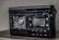 A damaged electronic device with a digital display showing the frequency 102.0 MHz. The surface of the device appears melted or burnt, with a rough texture around the front panel. The right side has several control buttons labeled with functions such as On/Off and Light.