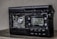 A damaged electronic device with a digital display showing the frequency 102.0 MHz. The surface of the device appears melted or burnt, with a rough texture around the front panel. The right side has several control buttons labeled with functions such as On/Off and Light.