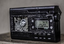 A damaged electronic device with a digital display showing the frequency 102.0 MHz. The surface of the device appears melted or burnt, with a rough texture around the front panel. The right side has several control buttons labeled with functions such as On/Off and Light.