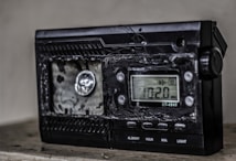 A damaged electronic device with a digital display showing the frequency 102.0 MHz. The surface of the device appears melted or burnt, with a rough texture around the front panel. The right side has several control buttons labeled with functions such as On/Off and Light.
