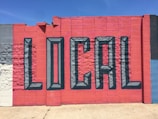 A vibrant mural painted on a concrete wall features the word 'LOCAL' in large, 3D-style letters. The wall is segmented into colorful blocks, primarily utilizing shades of red and blue, with a few sections in gray and white.
