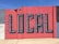 A vibrant mural painted on a concrete wall features the word 'LOCAL' in large, 3D-style letters. The wall is segmented into colorful blocks, primarily utilizing shades of red and blue, with a few sections in gray and white.