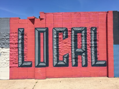A vibrant mural painted on a concrete wall features the word 'LOCAL' in large, 3D-style letters. The wall is segmented into colorful blocks, primarily utilizing shades of red and blue, with a few sections in gray and white.