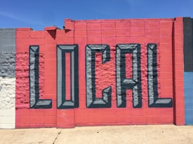 A vibrant mural painted on a concrete wall features the word 'LOCAL' in large, 3D-style letters. The wall is segmented into colorful blocks, primarily utilizing shades of red and blue, with a few sections in gray and white.