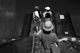 A black and white photograph of a person wearing a hat riding down an escalator, with two other people also on the escalator ahead, all carrying backpacks. The setting appears to be an indoor transit station or mall.