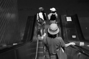 A black and white photograph of a person wearing a hat riding down an escalator, with two other people also on the escalator ahead, all carrying backpacks. The setting appears to be an indoor transit station or mall.