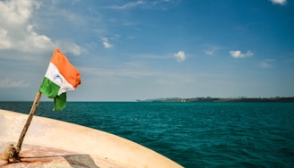 A boat deck with a small Indian flag fluttering in the wind, set against a backdrop of a vast, tranquil body of water and a distant shoreline under a mostly clear sky.