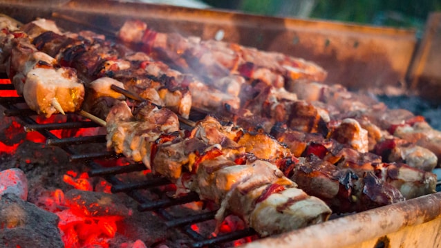 Skewers of grilled meat are sizzling over hot charcoal, with a warm glow of red and orange embers below. The meat appears to be well-cooked with slight charring visible.
