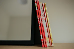 A set of six children's books with brightly colored spines, mostly red and yellow, leaning against the side of a black-framed mirror on a wooden surface.