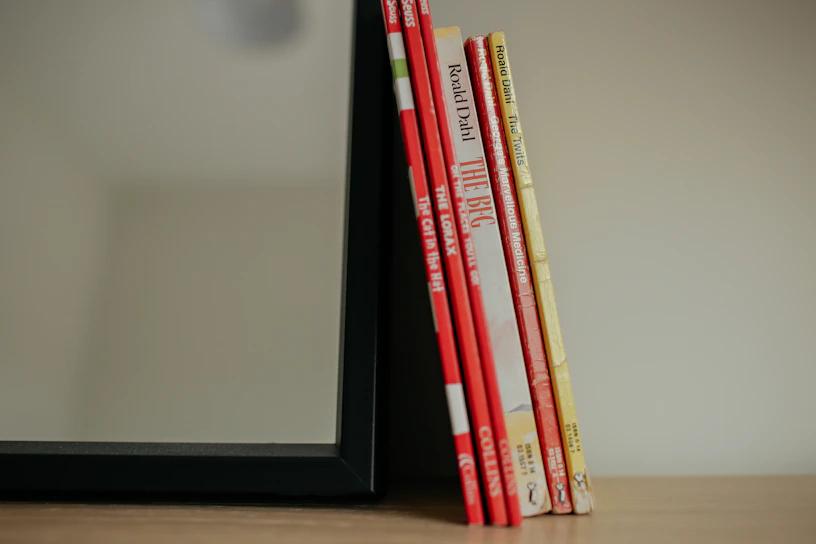 A set of six children's books with brightly colored spines, mostly red and yellow, leaning against the side of a black-framed mirror on a wooden surface.