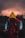 A person wearing a vibrant raincoat, featuring a vivid yellow hood, red body, and blue sleeves, stands on a shadowy surface. The background is dominated by dark, dramatic clouds with a subtle glowing effect, suggesting an impending storm or sunset. Silhouettes of trees frame the scene, adding to the overall mysterious and ominous atmosphere.