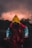 A person wearing a vibrant raincoat, featuring a vivid yellow hood, red body, and blue sleeves, stands on a shadowy surface. The background is dominated by dark, dramatic clouds with a subtle glowing effect, suggesting an impending storm or sunset. Silhouettes of trees frame the scene, adding to the overall mysterious and ominous atmosphere.
