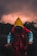 A person wearing a vibrant raincoat, featuring a vivid yellow hood, red body, and blue sleeves, stands on a shadowy surface. The background is dominated by dark, dramatic clouds with a subtle glowing effect, suggesting an impending storm or sunset. Silhouettes of trees frame the scene, adding to the overall mysterious and ominous atmosphere.