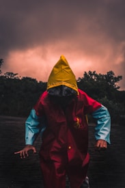 A person wearing a vibrant raincoat, featuring a vivid yellow hood, red body, and blue sleeves, stands on a shadowy surface. The background is dominated by dark, dramatic clouds with a subtle glowing effect, suggesting an impending storm or sunset. Silhouettes of trees frame the scene, adding to the overall mysterious and ominous atmosphere.