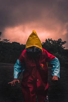 A person wearing a vibrant raincoat, featuring a vivid yellow hood, red body, and blue sleeves, stands on a shadowy surface. The background is dominated by dark, dramatic clouds with a subtle glowing effect, suggesting an impending storm or sunset. Silhouettes of trees frame the scene, adding to the overall mysterious and ominous atmosphere.