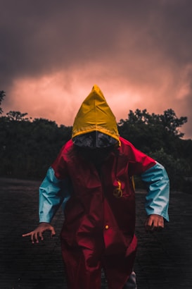 A person wearing a vibrant raincoat, featuring a vivid yellow hood, red body, and blue sleeves, stands on a shadowy surface. The background is dominated by dark, dramatic clouds with a subtle glowing effect, suggesting an impending storm or sunset. Silhouettes of trees frame the scene, adding to the overall mysterious and ominous atmosphere.