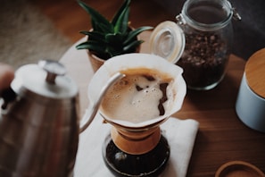 A pour-over coffee setup with a focus on the coffee being brewed. A kettle is pouring hot water over coffee grounds in a Chemex-style coffee maker. In the background, a jar of coffee beans and a potted plant add to the cozy atmosphere.