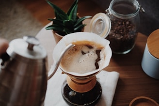 A pour-over coffee setup with a focus on the coffee being brewed. A kettle is pouring hot water over coffee grounds in a Chemex-style coffee maker. In the background, a jar of coffee beans and a potted plant add to the cozy atmosphere.