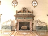 An ornate marble fireplace adorned with intricate carvings and sculptures. Flanking the fireplace are two reclining marble statues on green marble bases. Above the fireplace are three busts set within decorative niches.