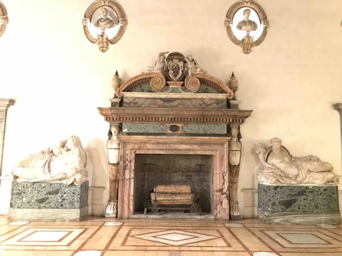 An ornate marble fireplace adorned with intricate carvings and sculptures. Flanking the fireplace are two reclining marble statues on green marble bases. Above the fireplace are three busts set within decorative niches.