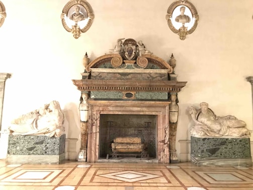An ornate marble fireplace adorned with intricate carvings and sculptures. Flanking the fireplace are two reclining marble statues on green marble bases. Above the fireplace are three busts set within decorative niches.