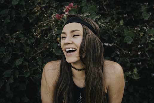 A person with long brown hair and a black headband is laughing joyfully against a backdrop of lush green foliage. The individual is wearing a black top and a choker necklace, with their eyes closed and a wide smile that conveys a sense of happiness and contentment.