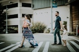A family of four crosses a street on a crosswalk. The mother is carrying a baby while wearing a blue floral dress. The father, dressed in a dark shirt and jeans, holds hands with a young girl in a white dress. Background features include a parking garage and some greenery with orange flowers.