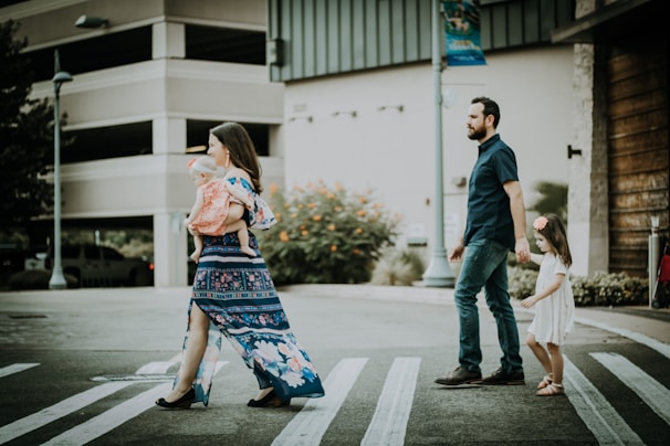 A family of four crosses a street on a crosswalk. The mother is carrying a baby while wearing a blue floral dress. The father, dressed in a dark shirt and jeans, holds hands with a young girl in a white dress. Background features include a parking garage and some greenery with orange flowers.