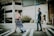 A family of four crosses a street on a crosswalk. The mother is carrying a baby while wearing a blue floral dress. The father, dressed in a dark shirt and jeans, holds hands with a young girl in a white dress. Background features include a parking garage and some greenery with orange flowers.