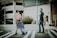 A family of four crosses a street on a crosswalk. The mother is carrying a baby while wearing a blue floral dress. The father, dressed in a dark shirt and jeans, holds hands with a young girl in a white dress. Background features include a parking garage and some greenery with orange flowers.
