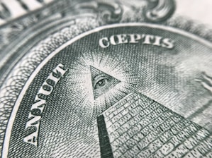 Close-up of a detailed engraving featuring a pyramid with an eye at its peak. This symbol is surrounded by Latin text 'Annuit Coeptis'. The intricate lines and patterns are rendered in shades of green, typical of paper currency.