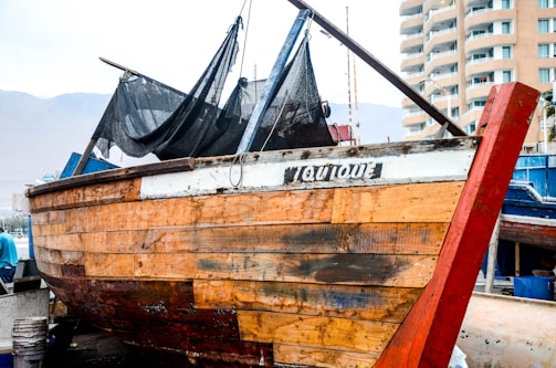 A wooden fishing boat is displayed in a dock area. The boat is weathered and has a mix of natural wood and painted surfaces. The name 'IQUIQUE' is painted on the side. In the background, there are modern residential buildings and a mountainous landscape under a cloudy sky.