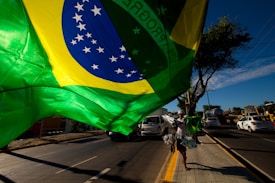 A large Brazilian flag is prominently displayed on a street, with a sunny sky in the background. There is a vendor walking along the sidewalk carrying more flags and other items. Cars are visible on the road, along with trees lining the street.