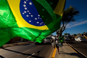 A large Brazilian flag is prominently displayed on a street, with a sunny sky in the background. There is a vendor walking along the sidewalk carrying more flags and other items. Cars are visible on the road, along with trees lining the street.