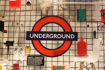 An array of various objects is displayed against a grid wall, centered around a prominent red circle with a blue bar across it displaying the word 'UNDERGROUND'. Items include a red bucket, a mop, a pair of scissors, a camera, white sneakers, a red hot water bottle, and a white chair. The setup has a modern, eclectic feel, incorporating items of different categories and functions.