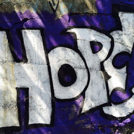 The image features a graffiti artwork on a rough, textured wall. The word 'Hope' is prominently displayed in large, bold letters. The letters are painted in white with black outlines and are set against a background of deep purple. Shadows and light variations add depth and dimension to the piece.