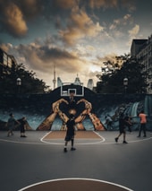 A group of people are playing basketball on an outdoor court with a large mural depicting hands reaching up toward a basketball hoop. The sky is overcast with dramatic clouds, and the city skyline, including a distinct tower, is visible in the background.