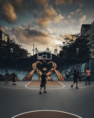 A group of people are playing basketball on an outdoor court with a large mural depicting hands reaching up toward a basketball hoop. The sky is overcast with dramatic clouds, and the city skyline, including a distinct tower, is visible in the background.