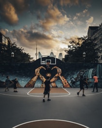 A group of people are playing basketball on an outdoor court with a large mural depicting hands reaching up toward a basketball hoop. The sky is overcast with dramatic clouds, and the city skyline, including a distinct tower, is visible in the background.