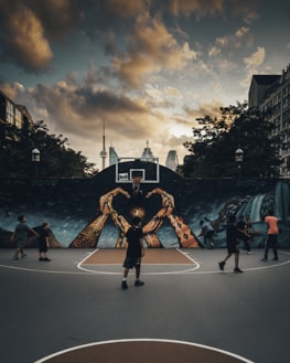 A group of people are playing basketball on an outdoor court with a large mural depicting hands reaching up toward a basketball hoop. The sky is overcast with dramatic clouds, and the city skyline, including a distinct tower, is visible in the background.