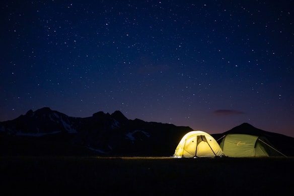 Two tents are illuminated at night under a starry sky, with mountains silhouetted in the background. One tent glows warmly with a light inside, providing a contrast to the deep blue and black of the sky and mountains.