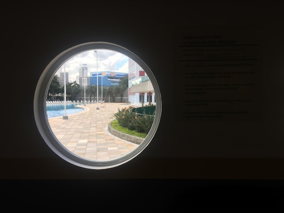 A circular window frames a view of an outdoor area with a swimming pool and lounge chairs. There are high-rise buildings visible in the background, along with some palm trees and other greenery. The pool is surrounded by a paved area and there are some poles visible, possibly for lighting or flags.