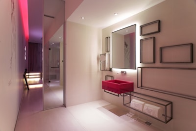 A modern bathroom with a sleek and minimalist design. It features a bold red sink mounted onto the wall below a large mirror that has a backlight. The wall next to the sink is decorated with various rectangular metal frame pieces of different sizes. There is a shelf below the sink holding rolled white towels. The room has a subtle and elegant lighting with a notable red accent light on the ceiling extending through the hallway. The floor is covered in light-colored tiles and the overall space has a contemporary and artistic feel with smooth surfaces and clean lines.