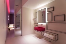 A modern bathroom with a sleek and minimalist design. It features a bold red sink mounted onto the wall below a large mirror that has a backlight. The wall next to the sink is decorated with various rectangular metal frame pieces of different sizes. There is a shelf below the sink holding rolled white towels. The room has a subtle and elegant lighting with a notable red accent light on the ceiling extending through the hallway. The floor is covered in light-colored tiles and the overall space has a contemporary and artistic feel with smooth surfaces and clean lines.